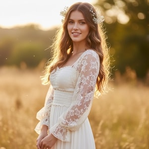 May include: A woman wearing a white lace dress with long sleeves and a square neckline, standing in a field. The dress has a fitted bodice and a flowing skirt. She wears a floral crown in her hair. The background is a blurred field of golden grass.