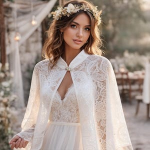 May include: A bride wearing a white lace wedding dress and cape with a floral crown. The dress features a V-neckline and a delicate lace overlay. The cape is fastened with a decorative clasp.