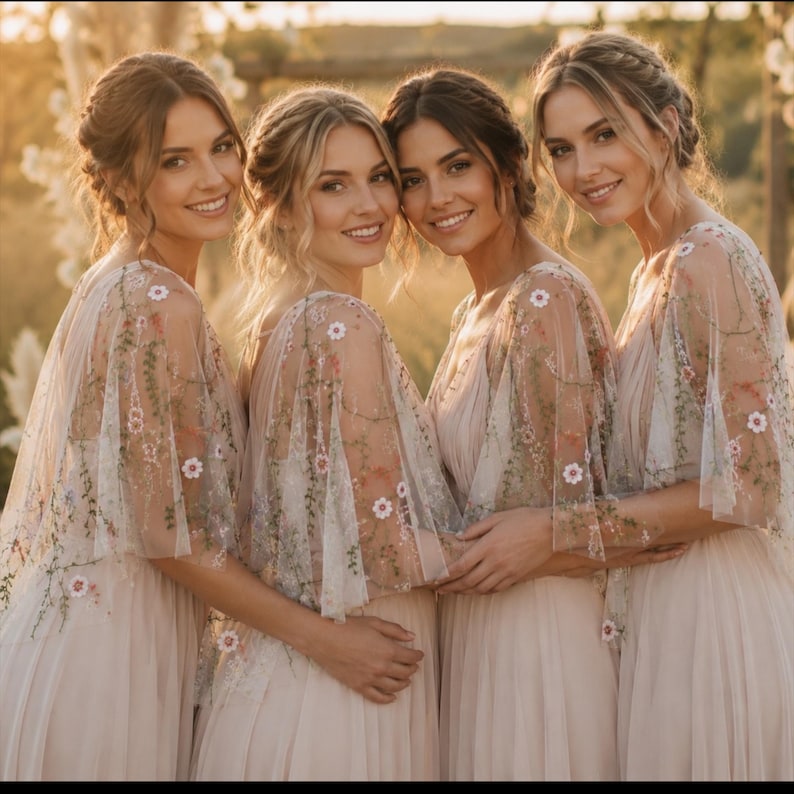 May include: Four individuals wearing matching floor-length, light beige dresses with sheer, flowing cap sleeves. The sleeves feature delicate, multi-colored floral embroidery with small white, pink, and green botanical patterns on a tulle fabric.