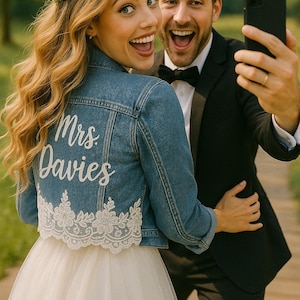 May include: A bride and groom take a selfie. The bride wears a denim jacket with "Mrs. Davies" in white script and a floral lace hem. She also wears a flower crown and a white wedding dress. The groom wears a black tuxedo and bow tie.