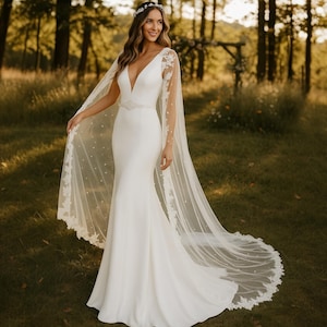 May include: A white wedding dress with a V-neckline and a flowing mermaid silhouette. The dress is paired with a sheer cape adorned with lace and pearl details. The bride is wearing a floral crown and a delicate belt.