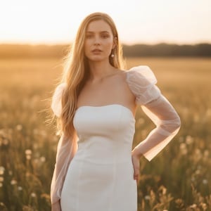 May include: A white strapless wedding dress with sheer, puffy sleeves. The dress is form-fitting and the sleeves are long and sheer. The background is a field of wildflowers at sunset.
