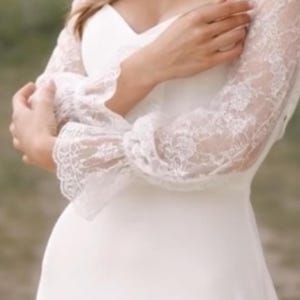 May include: A white wedding dress with long sleeves. The sleeves are made of delicate lace, and the dress has a simple, elegant design. The dress is a classic choice for a bride.