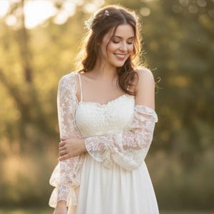 May include: A woman wearing a white wedding dress with spaghetti straps and a flowing skirt. The dress is paired with a delicate lace bolero with long sleeves. The background is a soft, blurred outdoor scene with natural light.
