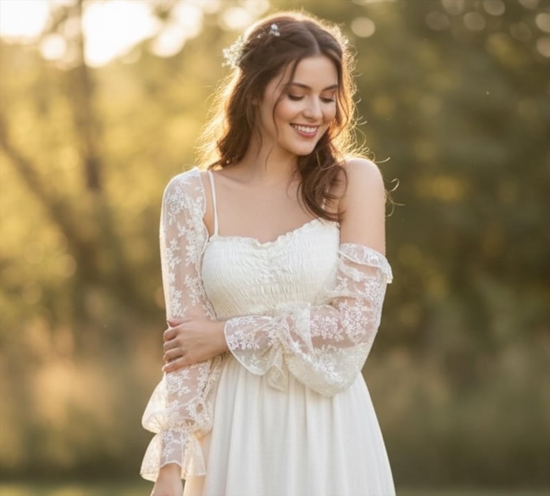 May include: A woman wearing a white wedding dress with spaghetti straps and a flowing skirt. The dress is paired with a delicate lace bolero with long sleeves. The background is a soft, blurred outdoor scene with natural light.