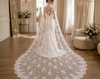 May include: A floor-length white wedding dress featuring a deep scoop back and a long, flowing lace cape veil. The veil is adorned with intricate floral embroidery and scalloped edges that extend across the floor in a wide, circular train.