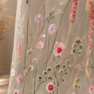 May include: A sheer, light-colored fabric featuring delicate embroidery of wildflowers in shades of pink, green, and white. The design includes tall, slender stems and various floral blooms scattered across the translucent material surface.