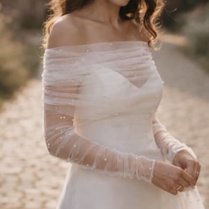Puede incluir: Un vestido blanco con hombros descubiertos que presenta mangas de tul transparente fruncido y una superposici&oacute;n en el corpi&ntilde;o adornada con peque&ntilde;as perlas blancas dispersas. La prenda tiene un escote coraz&oacute;n y mangas largas ajustadas.