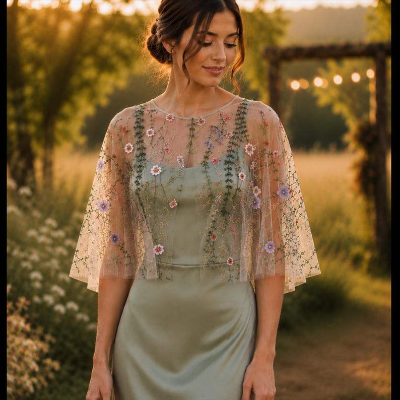 May include: A sage green satin dress featuring a sheer, short-sleeved overlay cape. The cape is made of fine mesh fabric adorned with delicate, multi-colored floral embroidery in shades of pink, purple, and green, creating a botanical pattern.