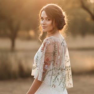 May include: A sheer white bridal capelet featuring delicate multi-colored floral embroidery. The lightweight tulle fabric drapes over the shoulders, showcasing intricate botanical patterns in shades of green, red, and pink across the back and sleeves.