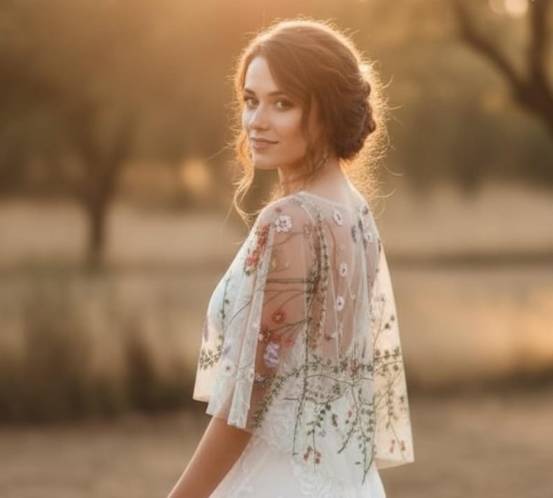 May include: A sheer white bridal capelet featuring delicate multi-colored floral embroidery. The lightweight tulle fabric drapes over the shoulders, showcasing intricate botanical patterns in shades of green, red, and pink across the back and sleeves.