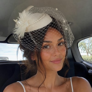 May include: A cream-colored pillbox hat featuring a delicate birdcage veil, adorned with a cluster of white feathers, a large central pearl, and a string of small pearls along the base. The netting is embellished with tiny white accents.
