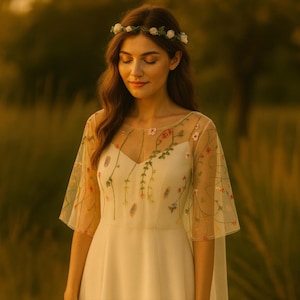 May include: A white dress featuring a sheer overlay with delicate floral embroidery on the bodice and sleeves, paired with a matching white floral crown. The dress has a V-neckline and flowing sleeves, creating a soft and elegant appearance.