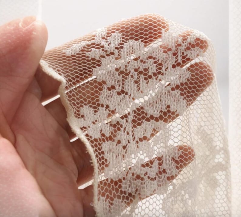 May include: Close-up of ivory lace fabric with a delicate floral pattern. The lace is held up, allowing light to pass through the intricate design. The fabric has a soft, textured appearance.