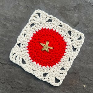 May include: A square crochet coaster featuring a circular bright red center with a small, embroidered gold-colored star at its core, surrounded by an off-white, open-work lacy border with a repeating shell-like pattern along the edges.