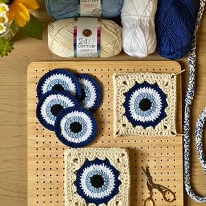 May include: A bamboo blocking board with crochet projects including three circular evil eye coasters and two square granny squares. Included are three skeins of 24/7 Cotton yarn in light blue, cream, and navy, plus a pair of gold stork scissors.
