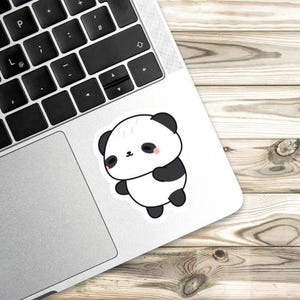 K&ouml;nnte beinhalten: Ein wei&szlig;er Aufkleber mit einem Cartoon-Panda-B&auml;ren mit schwarzen Ohren, Armen und F&uuml;&szlig;en. Der Panda hat ein kleines rosa Rouge auf den Wangen und befindet sich auf einem Laptop. Der Hintergrund ist eine Holzoberfl&auml;che.