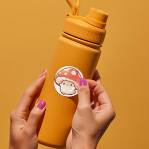 K&ouml;nnte beinhalten: Eine senfgelbe Wasserflasche mit passendem Deckel und einem wei&szlig;en Aufkleber mit einem Cartoon-Pilz mit roter und wei&szlig;er Kappe. Die Flasche wird von H&auml;nden mit lila Nagellack vor einem gelben Hintergrund gehalten.