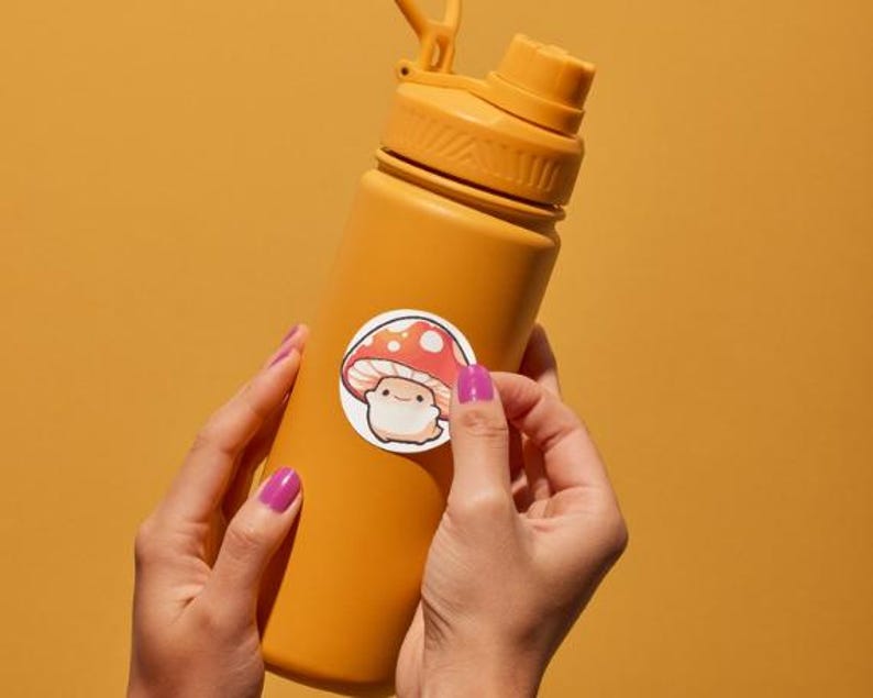 K&ouml;nnte beinhalten: Eine senfgelbe Wasserflasche mit passendem Deckel und einem wei&szlig;en Aufkleber mit einem Cartoon-Pilz mit roter und wei&szlig;er Kappe. Die Flasche wird von H&auml;nden mit lila Nagellack vor einem gelben Hintergrund gehalten.