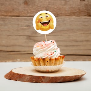 K&ouml;nnte beinhalten: Ein Cupcake mit wei&szlig;em Zuckerguss und bunten Streuseln, gekr&ouml;nt von einem runden Emoji-Gesicht auf einem Holzstab. Der Cupcake steht auf einer Holzscheibe, mit einem Holz-Hintergrund.