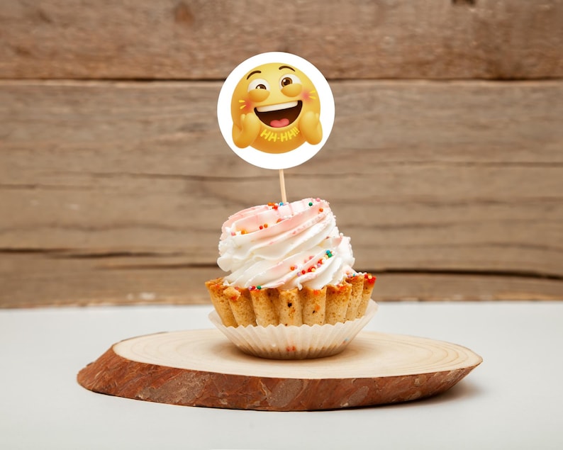 K&ouml;nnte beinhalten: Ein Cupcake mit wei&szlig;em Zuckerguss und bunten Streuseln, gekr&ouml;nt von einem runden Emoji-Gesicht auf einem Holzstab. Der Cupcake steht auf einer Holzscheibe, mit einem Holz-Hintergrund.
