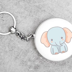 K&ouml;nnte beinhalten: Ein runder, wei&szlig;er Schl&uuml;sselanh&auml;nger mit einer Cartoon-Elefanten-Illustration. Der Elefant ist hellblau mit pfirsichfarbenen Ohren und rosigen Wangen. Der Schl&uuml;sselanh&auml;nger hat einen silbernen Ring und eine Kette.