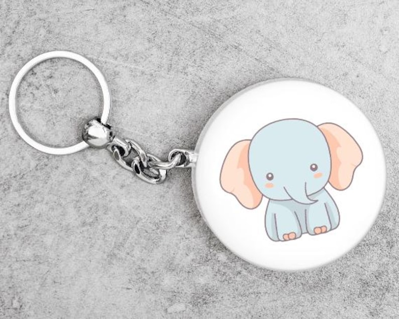 K&ouml;nnte beinhalten: Ein runder, wei&szlig;er Schl&uuml;sselanh&auml;nger mit einer Cartoon-Elefanten-Illustration. Der Elefant ist hellblau mit pfirsichfarbenen Ohren und rosigen Wangen. Der Schl&uuml;sselanh&auml;nger hat einen silbernen Ring und eine Kette.