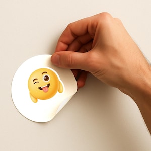 K&ouml;nnte beinhalten: Ein runder, wei&szlig;er Aufkleber mit einem gelben Emoji, das ein zwinkerndes Auge und eine herausgestreckte Zunge zeigt. Das Emoji hat einen 3D-Effekt und einen l&auml;chelnden Ausdruck. Der Aufkleber wird von einer Hand gehalten.