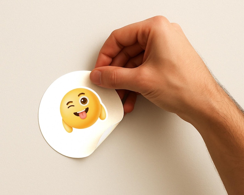 K&ouml;nnte beinhalten: Ein runder, wei&szlig;er Aufkleber mit einem gelben Emoji, das ein zwinkerndes Auge und eine herausgestreckte Zunge zeigt. Das Emoji hat einen 3D-Effekt und einen l&auml;chelnden Ausdruck. Der Aufkleber wird von einer Hand gehalten.