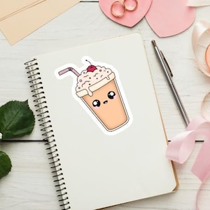 K&ouml;nnte beinhalten: Ein Aufkleber eines Cartoon-Milchshakes mit Kirsche, Strohhalm und einem niedlichen Gesicht. Der Milchshake ist hellbraun mit wei&szlig;er Schlagsahne. Der Aufkleber befindet sich auf einem wei&szlig;en Spiralnotizbuch.