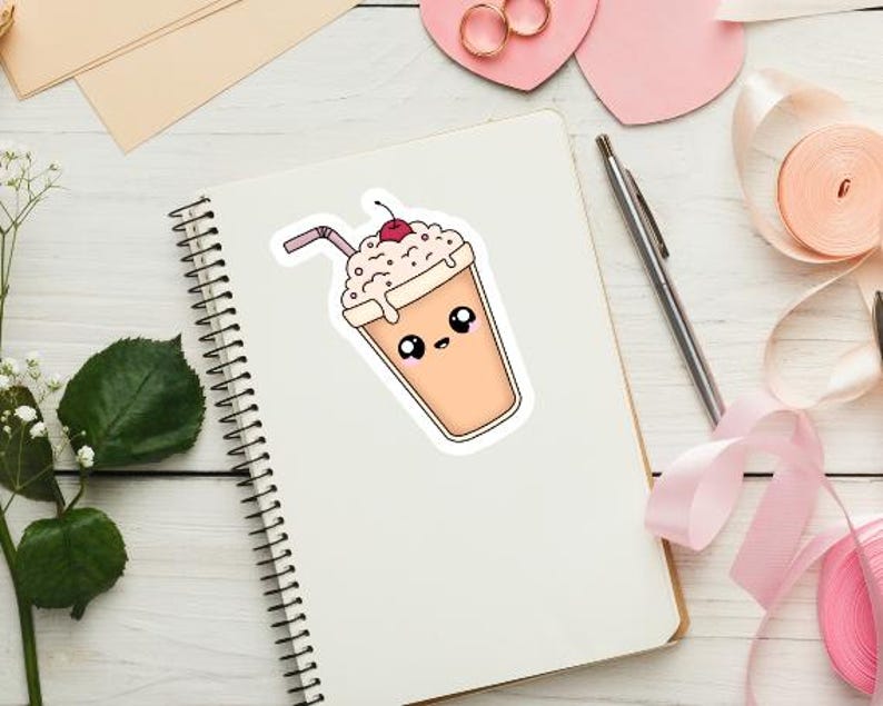 K&ouml;nnte beinhalten: Ein Aufkleber eines Cartoon-Milchshakes mit Kirsche, Strohhalm und einem niedlichen Gesicht. Der Milchshake ist hellbraun mit wei&szlig;er Schlagsahne. Der Aufkleber befindet sich auf einem wei&szlig;en Spiralnotizbuch.