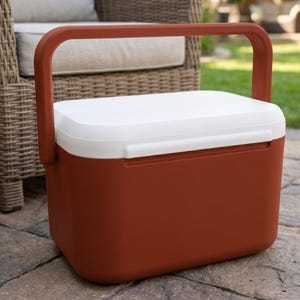 Peut inclure: Glacière rectangulaire avec un couvercle blanc et un corps de couleur rouille. La glacière a une poignée de couleur rouille assortie pour faciliter le transport. Conçue pour une utilisation en extérieur, elle est posée sur un dallage en pierre.