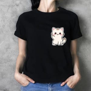 K&ouml;nnte beinhalten: Schwarzes T-Shirt mit einem niedlichen Cartoon-Katzen-Motiv auf der Vorderseite. Die Katze ist wei&szlig; mit braunen Akzenten und winkt. Das Shirt hat kurze &Auml;rmel und einen Rundhalsausschnitt.