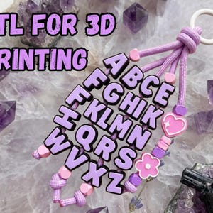 Peut inclure: Un porte-clés avec des lettres de l'alphabet violet clair disposées en rangées, attachées à des brins de paracorde lavande avec un anneau blanc. Le design inclut de petites perles en forme de cœur et de fleur roses. Le texte STL FOR 3D PRINTING est visible.