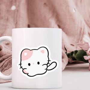 K&ouml;nnte beinhalten: Wei&szlig;e Keramik-Tasse mit einem Cartoon-Katzen-Aufkleber. Die Katze ist wei&szlig; mit einem rosa Ohr und einer Pfote, schwarz umrandet. Die Tasse hat einen wei&szlig;en Henkel und steht vor einem rosa Stoffhintergrund mit einer rosa Blume.