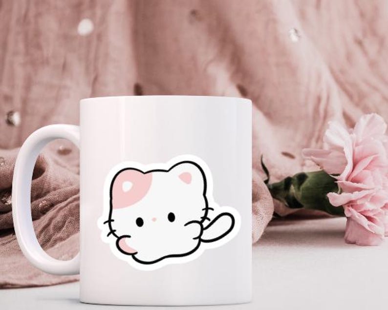 K&ouml;nnte beinhalten: Wei&szlig;e Keramik-Tasse mit einem Cartoon-Katzen-Aufkleber. Die Katze ist wei&szlig; mit einem rosa Ohr und einer Pfote, schwarz umrandet. Die Tasse hat einen wei&szlig;en Henkel und steht vor einem rosa Stoffhintergrund mit einer rosa Blume.
