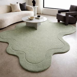 May include: A pale green, organically shaped rug with a distinctive wavy outline. The rug sits on a concrete floor, complemented by a cream sofa, a marble coffee table, and a dark grey armchair. The rug's edges feature a slightly darker border.