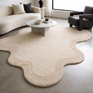 May include: A beige, organically shaped area rug with a wavy border. The rug is placed on a gray concrete floor, and is surrounded by furniture, including a sofa, armchair, and coffee table.