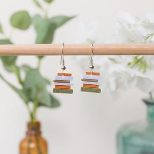 May include: Silver earrings, each with a miniature stack of books in green, white, orange, and brown. The earrings hang from a wooden dowel, with a blurred backdrop of green foliage and white flowers. Each book stack is approximately 1 cm high.