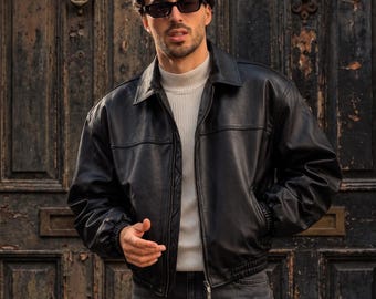 May include: A black leather jacket with a classic collar, front zipper closure, and elasticized cuffs and hem, worn over a white ribbed mock neck sweater and dark gray denim jeans, paired with black rectangular sunglasses.