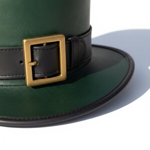 May include: A green top hat with a black brim and a black band featuring a gold-colored buckle. The hat is made of a smooth material and is set against a white background.