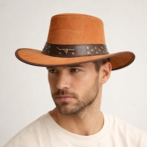 May include: A tan-colored cowboy hat with a wide brim and a cylindrical crown. The hat features a dark brown leather band adorned with rivets and a metal bull skull emblem. The hat is made of a suede-like material.