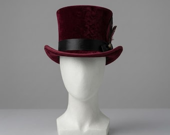 May include: A deep burgundy velvet top hat featuring a wide black satin ribbon band around the base of the crown. The hat is accented with a dark feather detail tucked into the side of the band, secured by a small metallic circular button.
