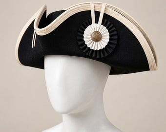 May include: A black tricorn hat featuring a cream-colored trim along the edges. The hat is adorned with a layered circular cockade consisting of black and white pleated fabric, centered with a metallic bronze-toned button detail.