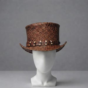 Puede incluir: Un sombrero de copa marrón con un patrón texturizado similar a la piel de reptil en toda la superficie. El sombrero presenta una banda decorativa alrededor de la base adornada con pequeños detalles claros en forma de diente sujetos por cordones oscuros.