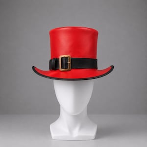 Puede incluir: Un sombrero de copa rojo brillante con un acabado liso y un ala ancha y curvada. El sombrero está acentuado con una banda negra similar al cuero que envuelve la base, asegurada por una hebilla rectangular de tono latón centrada al frente.