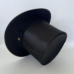 Puede incluir: Un sombrero de copa negro con ala ancha y corona cil&iacute;ndrica. El sombrero presenta una banda negra alrededor de la corona y una peque&ntilde;a correa asegurada con un bot&oacute;n dorado. El sombrero est&aacute; hecho de una tela oscura y lisa.