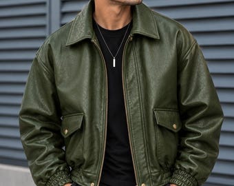 May include: An olive green bomber jacket with a textured finish, featuring a classic collar, a full-length front zipper, and two front flap pockets with gold-tone snap buttons. The jacket has ribbed cuffs and a ribbed hem, worn over a black shirt.