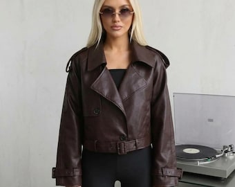 May include: A dark brown cropped faux leather jacket featuring a wide collar, long sleeves with buckled cuffs, and a matching belt at the waist. The jacket has a button closure, shoulder epaulets, and a smooth finish, worn over a black top.
