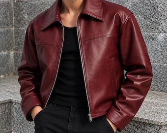 May include: A deep burgundy leather jacket featuring a classic pointed collar, long sleeves, and a full-length silver-toned metal zipper closure. The jacket is worn over a simple black tank top and paired with black trousers, showcasing a smooth finish.